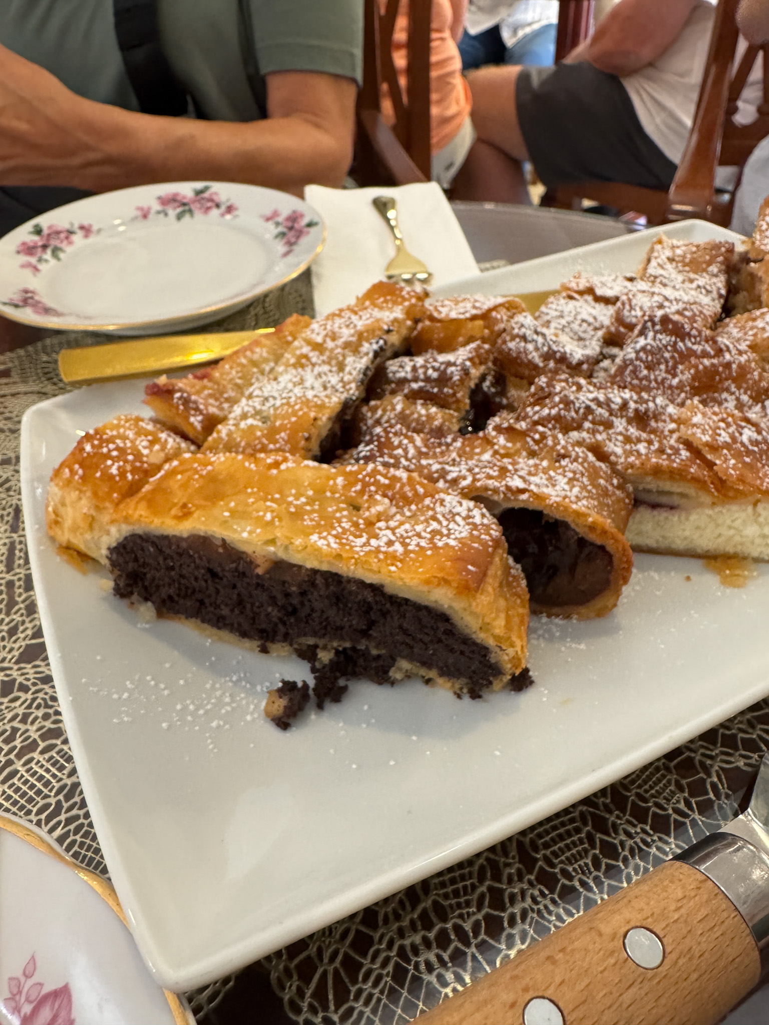 A sliced poppy seed Hungarian pastry dusted with powdered sugar on a white plate, with plates and cutlery nearby.