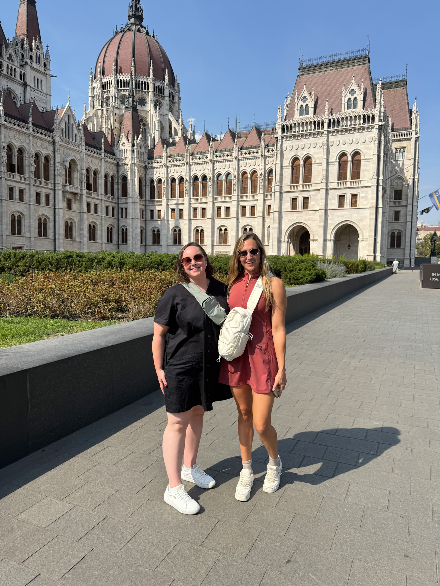 Two women posing and smiling in front of the Hungarian Parliament building on a sunny day.