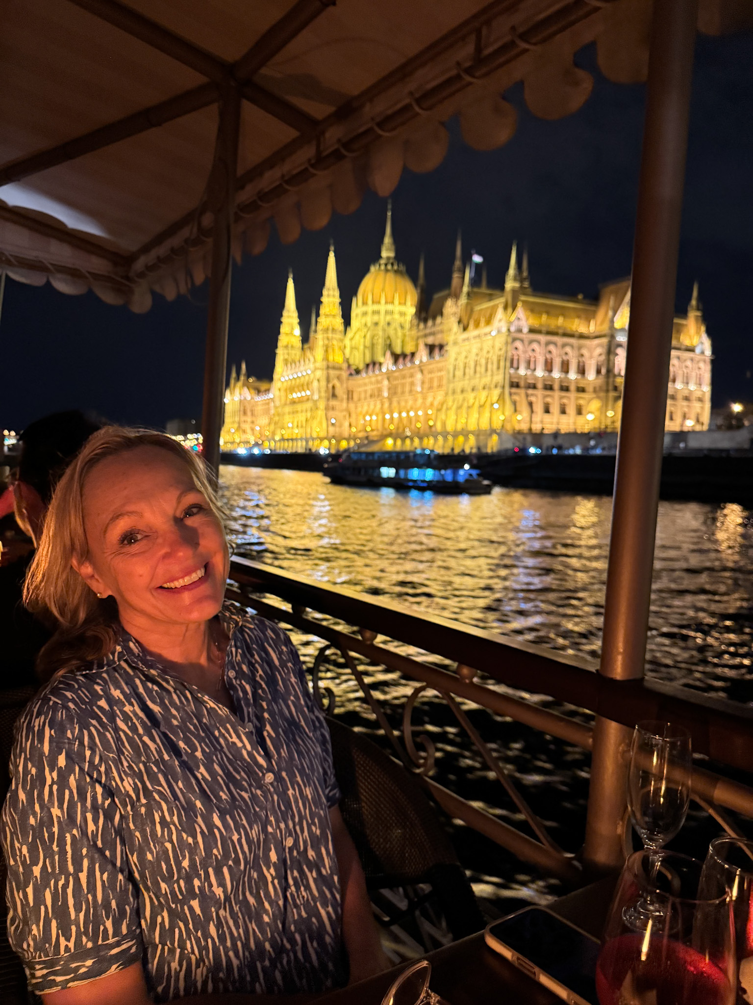 Smiling woman on a boat at night with the illuminated Hungarian Parliament building in the background.