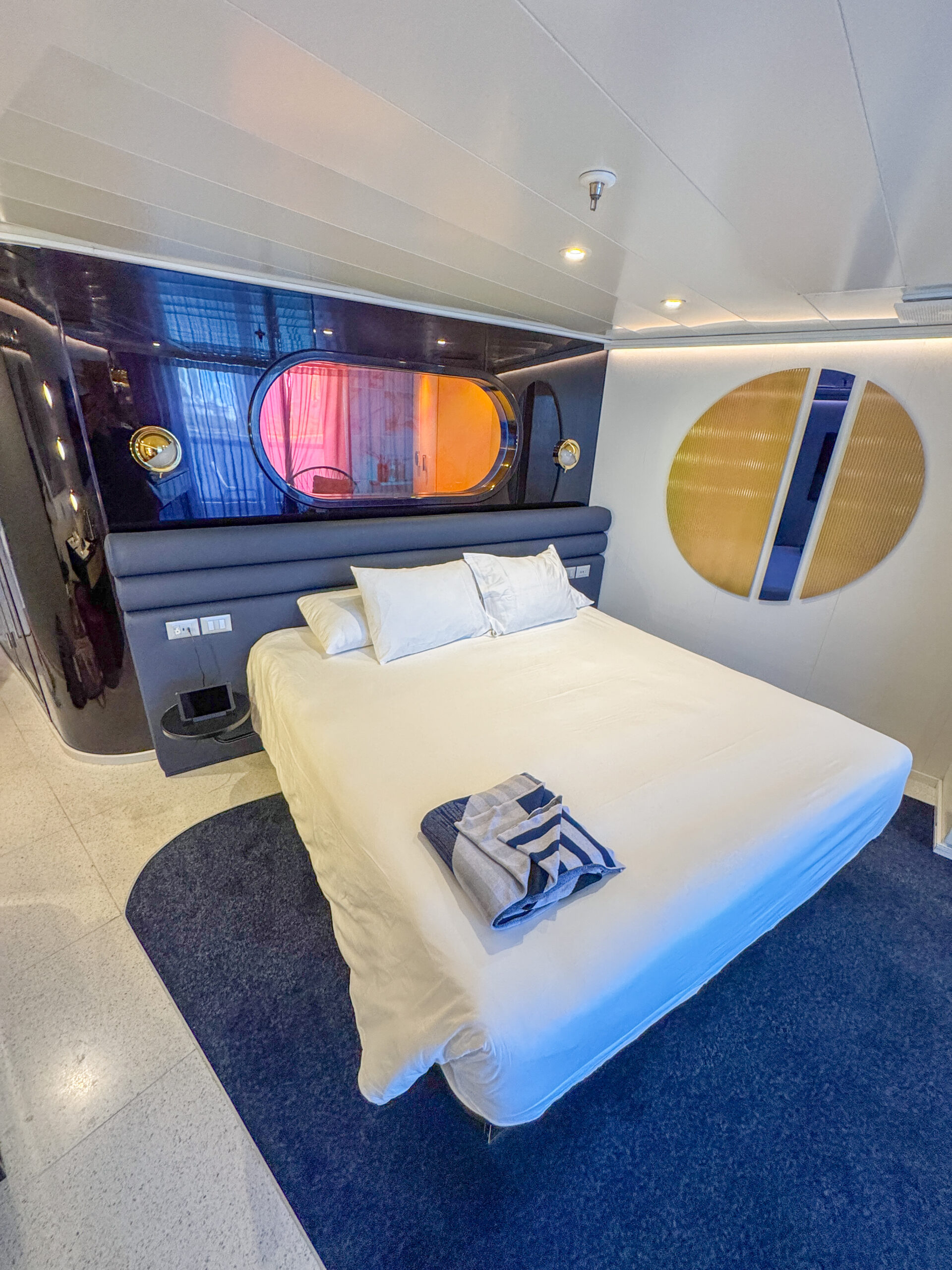Modern bedroom with a white bed, blue carpet, round window, and abstract wall art. Towel placed on the bed.