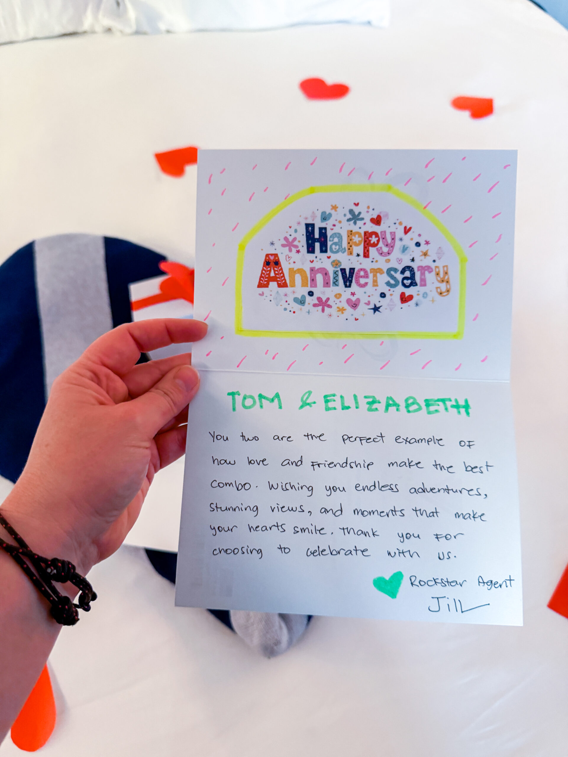 A hand holds an anniversary card with colorful writing above a bed decorated with red heart shapes.