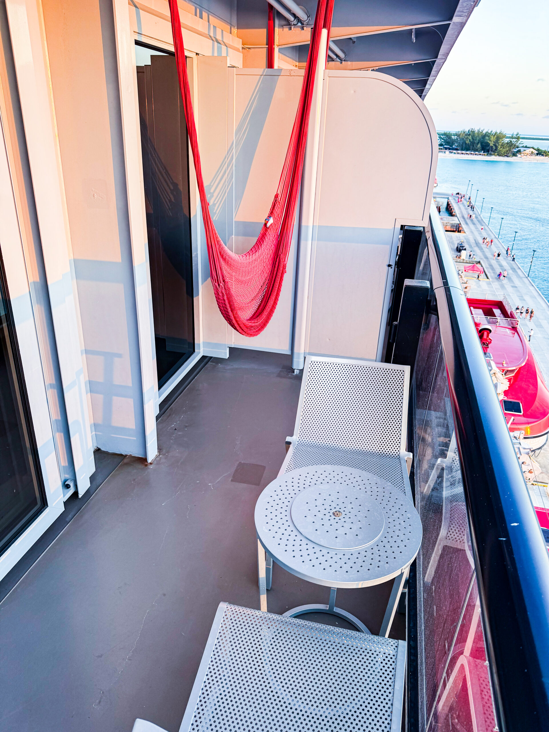 View from a modern cruise terminal lounge with floor-to-ceiling windows overlooking a docked Virgin Voyages cruise ship. Blue lounge chairs and a small table sit beside the glass, while the ship, pier, and harbor are visible under a bright blue sky.