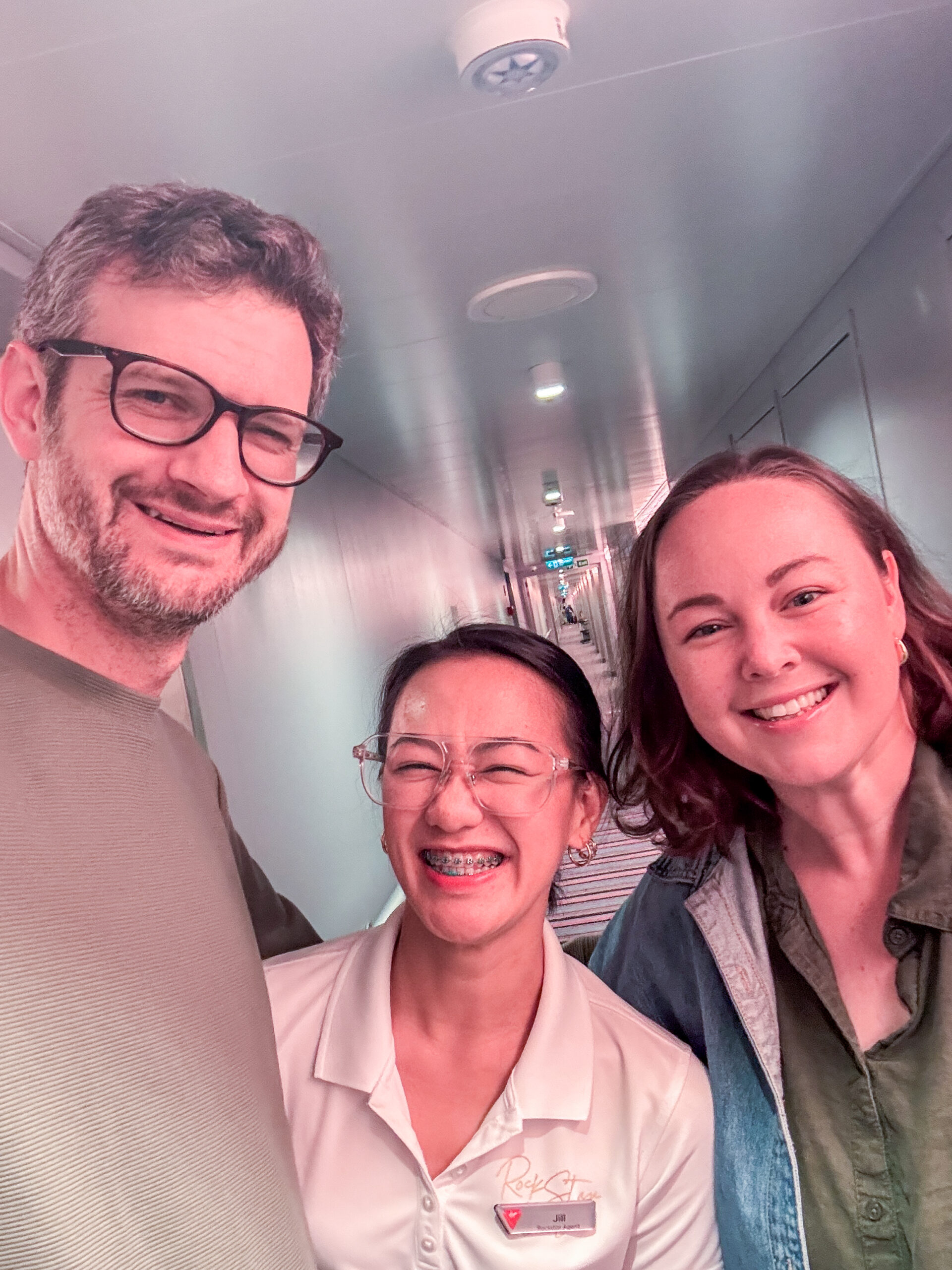 Three people smiling in a hallway; two are casually dressed, one wears a name tag and uniform shirt.