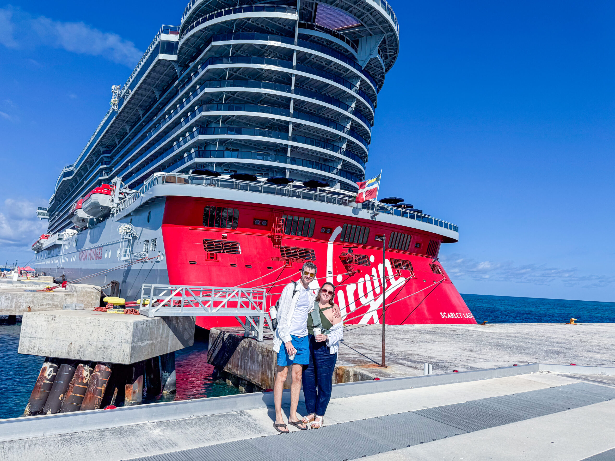 Couple standing on a cruise pier in front of the bright red stern of a Virgin Voyages cruise ship under a clear blue sky. The massive ship towers overhead while turquoise Caribbean waters surround the dock.