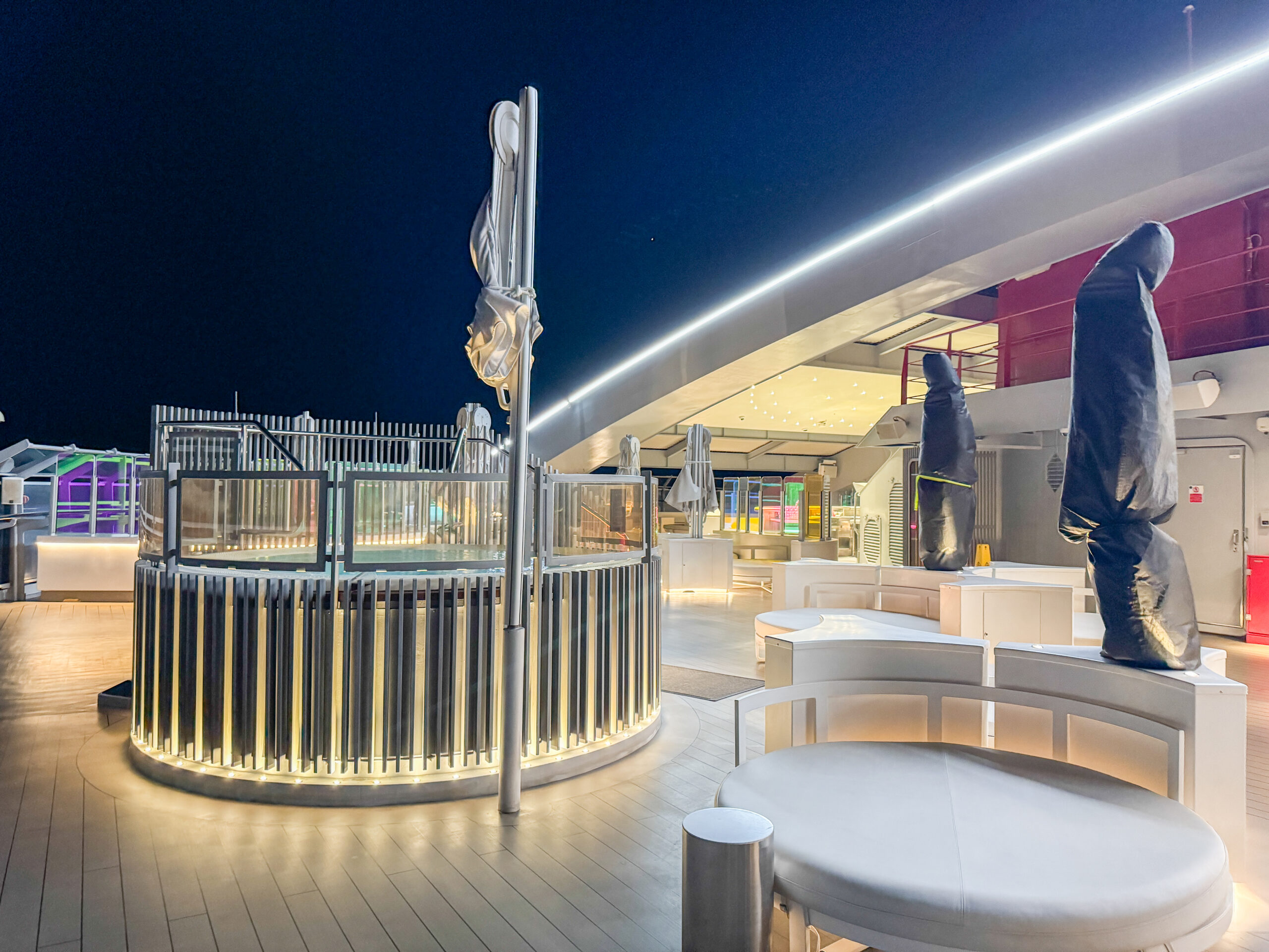 Modern cruise ship deck at night with illuminated seating and covered furniture under bright lights.