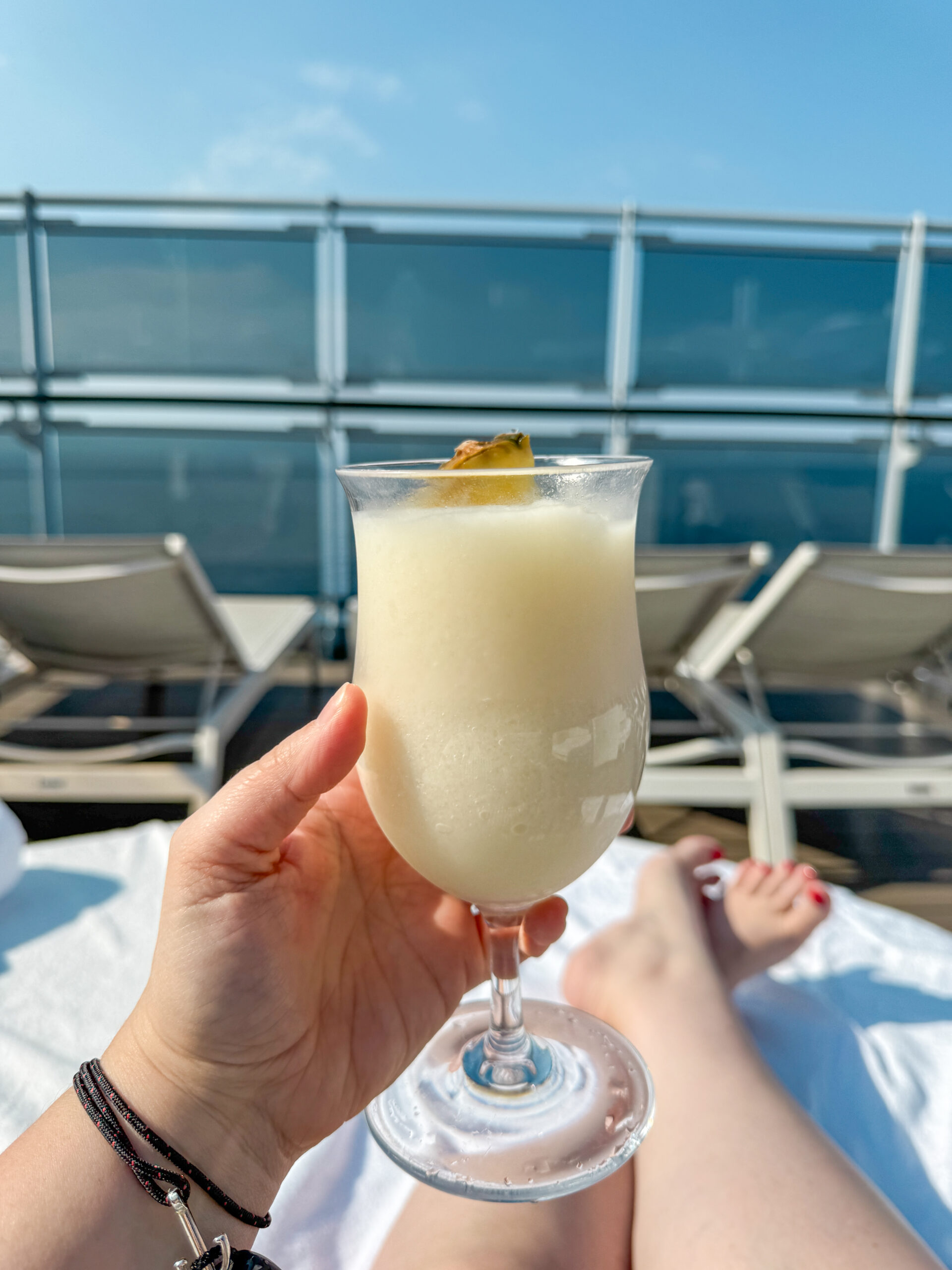 A hand holds a pina colada by a poolside with lounge chairs and blue sky in the background.