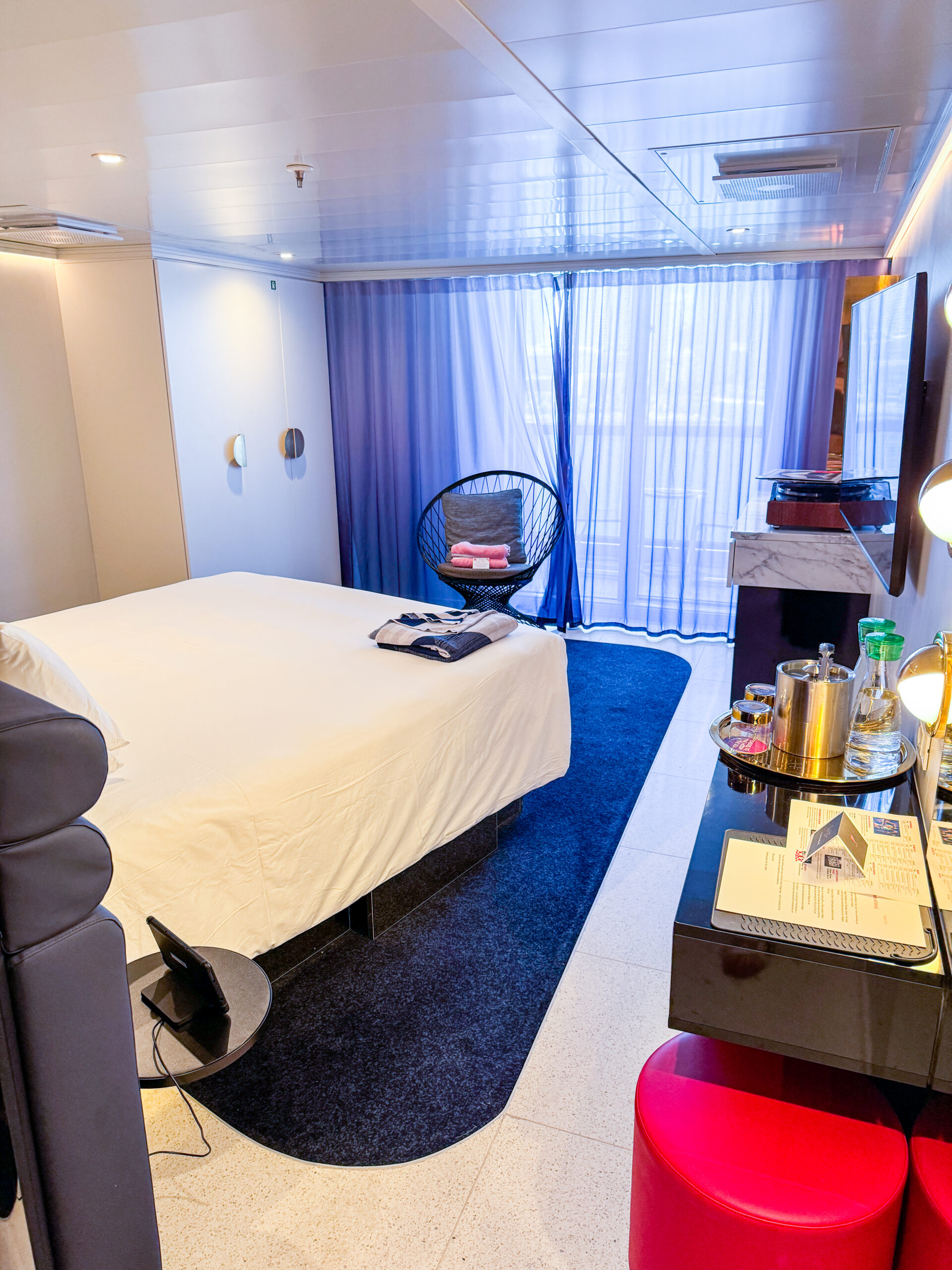 Modern bedroom with a white bed, blue carpet, vanity, curtains covering balcony area, mini bar.