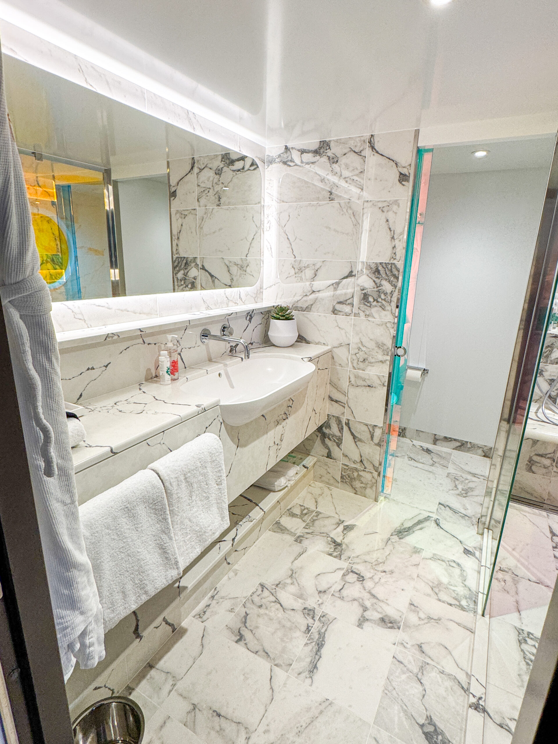 Modern bathroom with marble walls and floor, a sink, mirror, towels, plant, and toiletries on the counter.