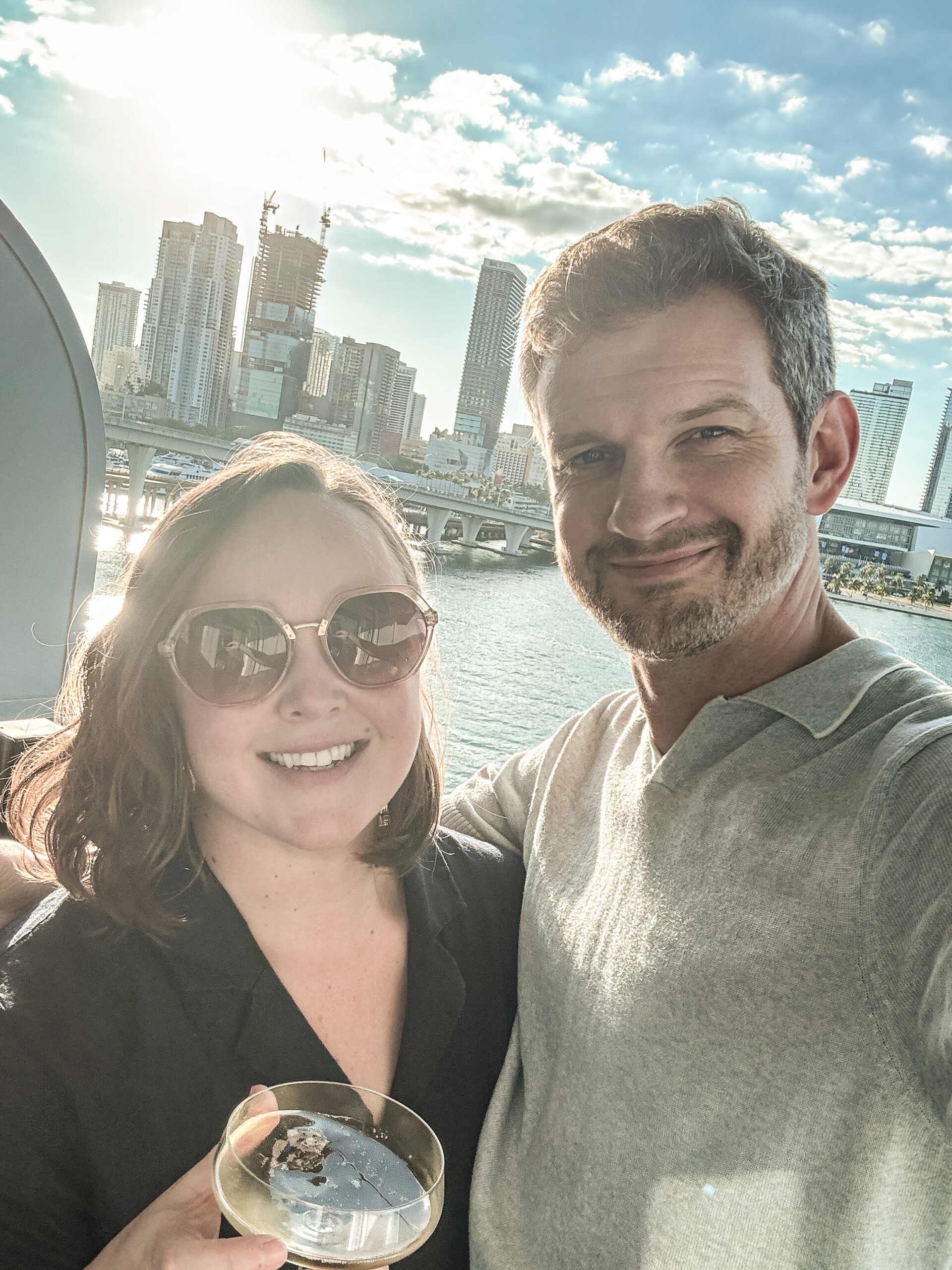 A smiling couple takes a selfie on a balcony overlooking a city skyline and water, with sunlight behind them.