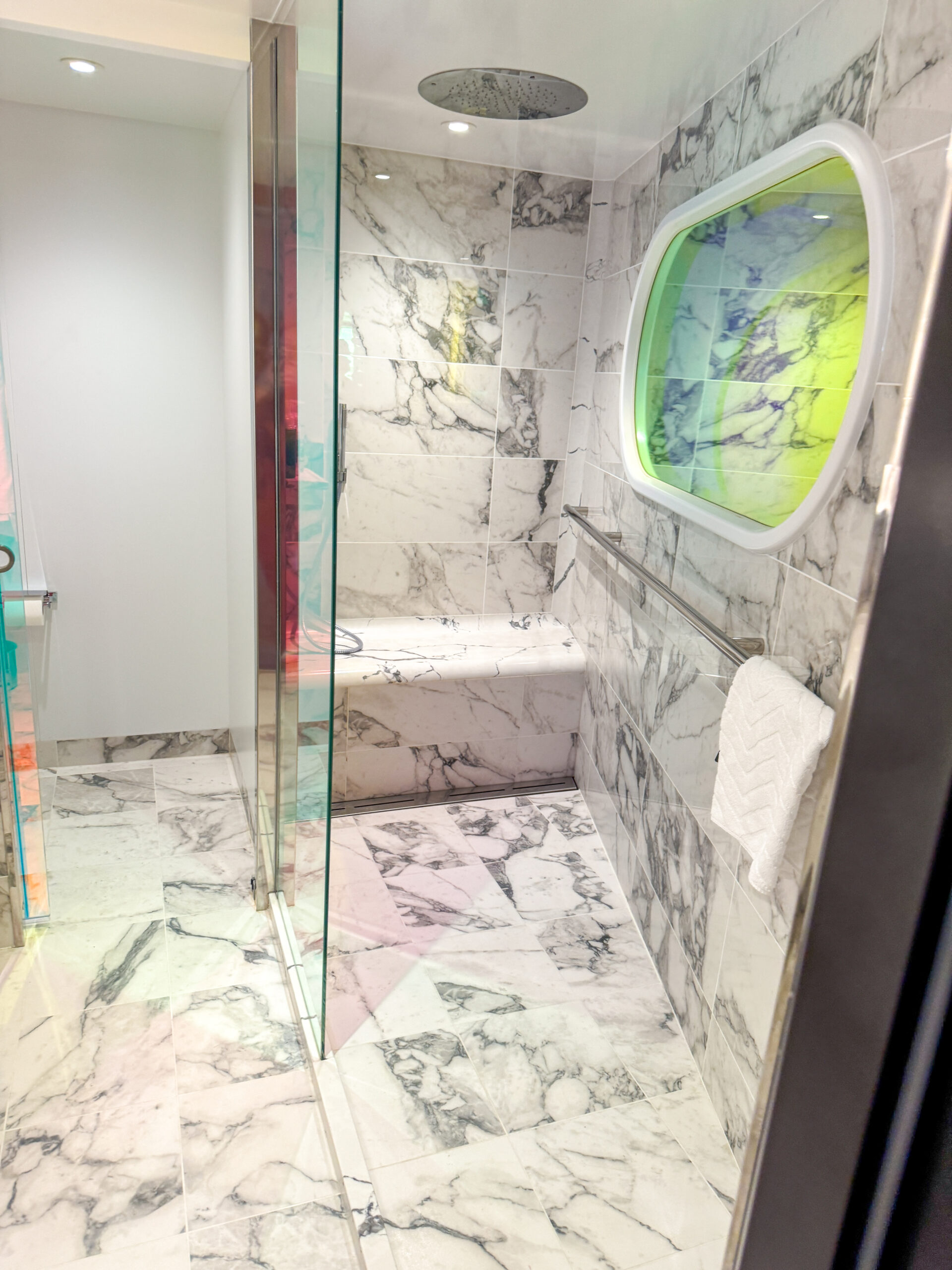 Marble-tiled shower with glass door, built-in bench, grab bar, and oval window with colorful glass.