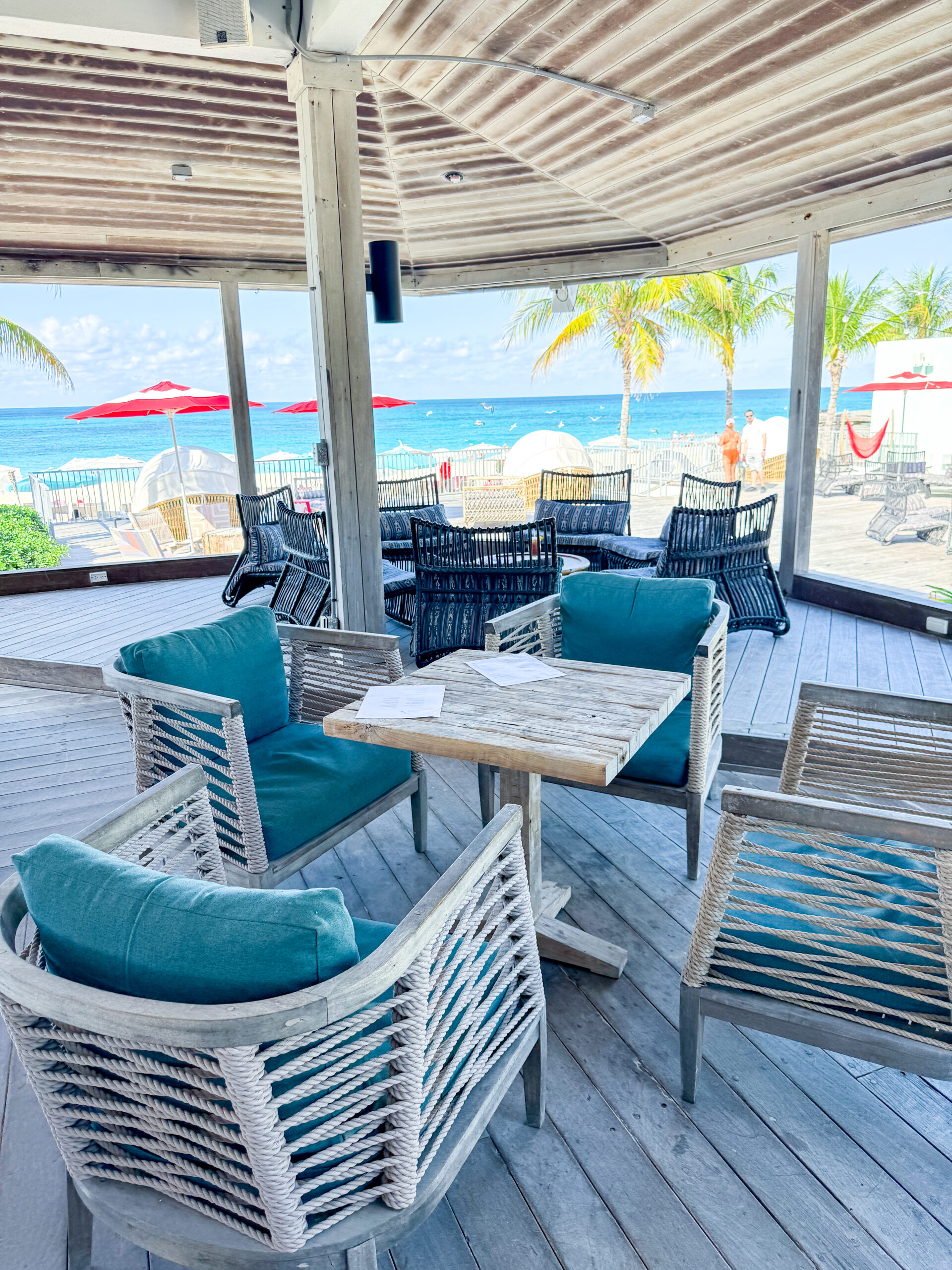 Covered beachfront lounge area with woven chairs and a rustic wooden table overlooking a turquoise ocean. Palm trees, red umbrellas, and cabanas line the deck outside, creating a relaxed tropical resort setting with sea views.